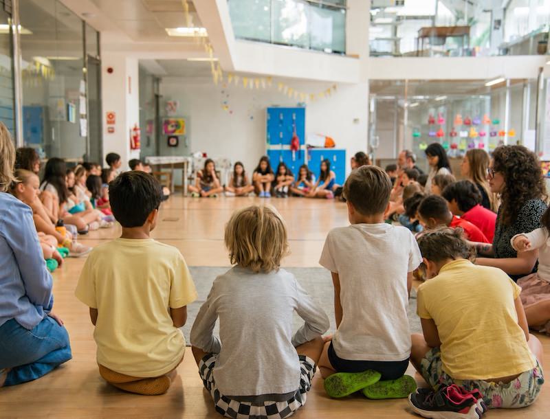 A large group of students and educators sitting together in a circle on the wooden floor during an assembly at the Second Body Montessori school in Valencia.
