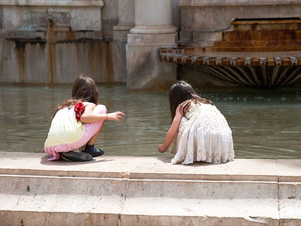 Two little girls crouching and playing with water at the edge of a monumental classic stone fountain.